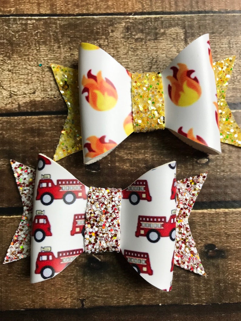May include: Two handmade hair bows with glitter accents. One bow features a white background with a red firetruck pattern. The other bow features a white background with a yellow flame pattern.