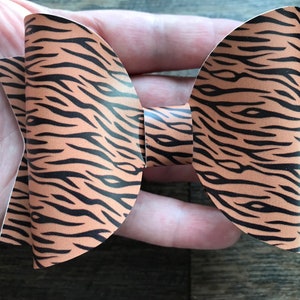May include: A handmade hair bow featuring a tiger stripe pattern. The bow is made of a tan material with black stripes. The bow is held in a person's hand.