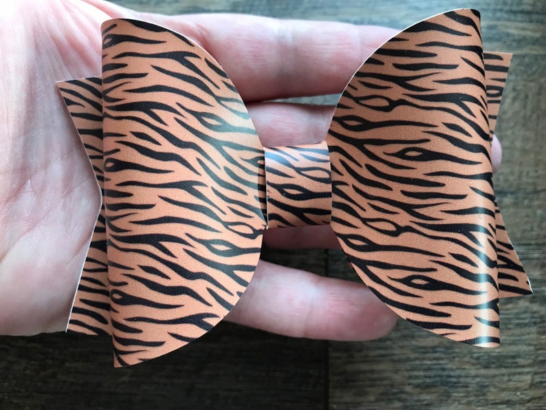 May include: A handmade hair bow featuring a tiger stripe pattern. The bow is made of a tan material with black stripes. The bow is held in a person's hand.