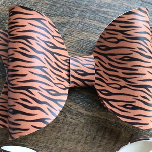May include: A large, handmade hair bow with a tiger stripe pattern in shades of brown and black. The bow is made of a textured material and is displayed on a wooden surface. Other bows with different patterns are partially visible.