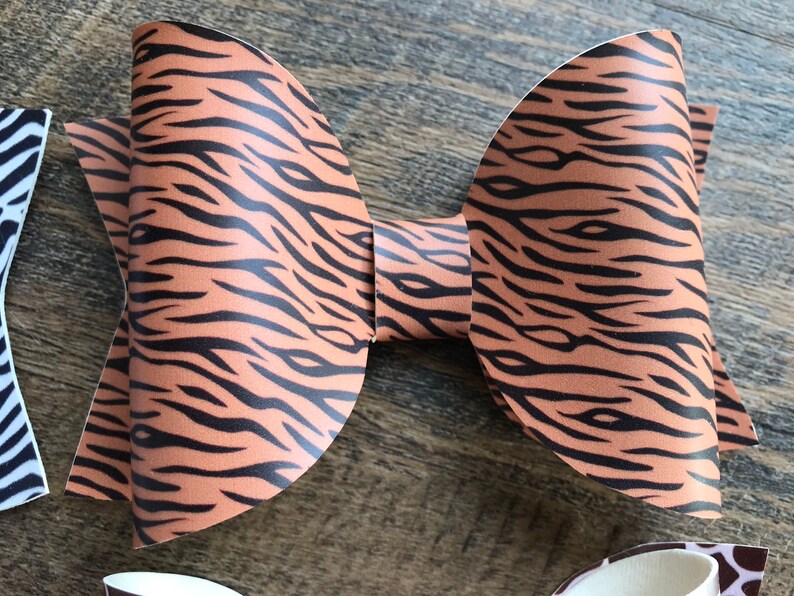 May include: A large, handmade hair bow with a tiger stripe pattern in shades of brown and black. The bow is made of a textured material and is displayed on a wooden surface. Other bows with different patterns are partially visible.