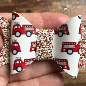 May include: A white and red glitter hair bow with a fire truck pattern. The bow is made of fabric and has a red glitter center.