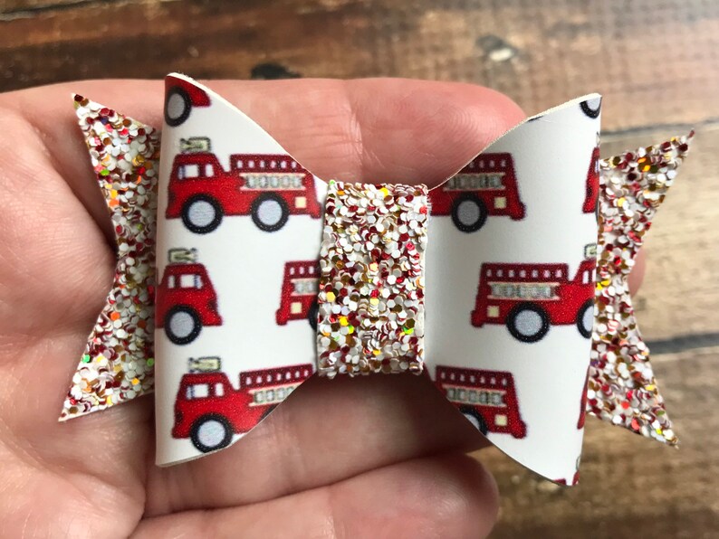 May include: A white and red glitter hair bow with a fire truck pattern. The bow is made of fabric and has a red glitter center.