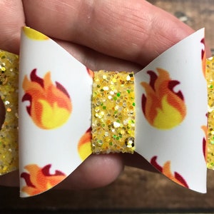 May include: A yellow glitter hair bow with a white bow center featuring a repeating pattern of red and yellow flames.