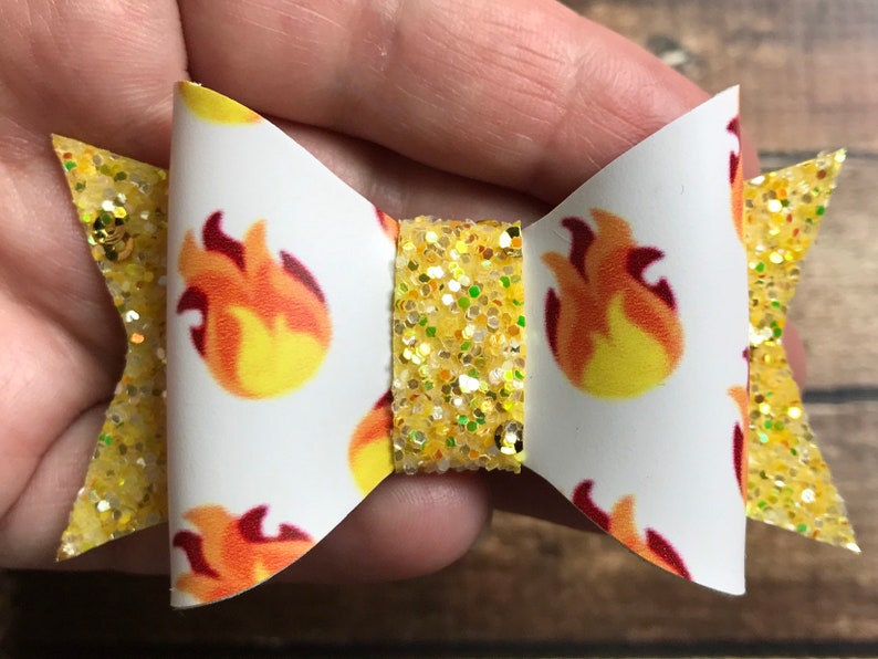 May include: A yellow glitter hair bow with a white bow center featuring a repeating pattern of red and yellow flames.