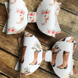 May include: Two white hair bows with different patterns. One bow has a white background with a repeated pattern of brown horses with pink flowers. The other bow has a white background with a repeated pattern of brown cowboy boots with pink flowers.