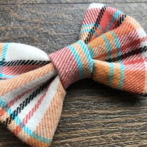 May include: A plaid bow tie in shades of orange, white, black, and blue. The bow tie is made of a soft, woven fabric.