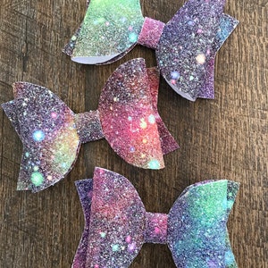 May include: Three sparkly galaxy-themed hair bows in shades of pink, purple, and green. Each bow has a glittery finish and a white backing.