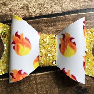 May include: A white bow with a yellow glitter center and a pattern of orange flames. The bow is made of fabric and has a white background.