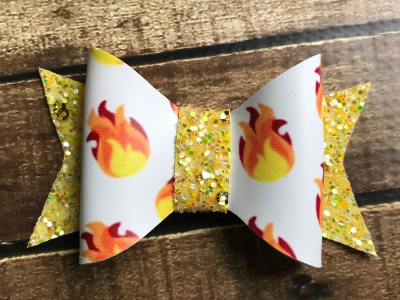 May include: A white bow with a yellow glitter center and a pattern of orange flames. The bow is made of fabric and has a white background.