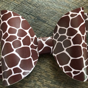 May include: A brown and white giraffe print bow. The bow is made of a smooth material and has a classic bow shape. The giraffe print features irregular brown shapes on a white background. The bow is a hair accessory.