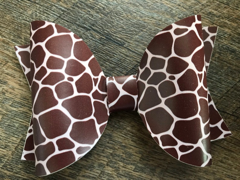 May include: A brown and white giraffe print bow. The bow is made of a smooth material and has a classic bow shape. The giraffe print features irregular brown shapes on a white background. The bow is a hair accessory.