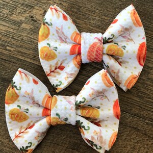 May include: Two white fabric hair bows with a pumpkin and leaf pattern. The bows are tied with a white ribbon.