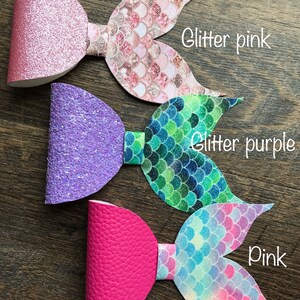 May include: Three mermaid tail hair bows with glitter bows. The bows are pink, purple, and a solid pink. The tails are pink with glitter scales, green with glitter scales, and blue with glitter scales.