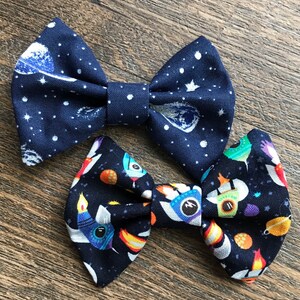 May include: Two navy blue bow ties with different space-themed patterns. One bow tie features a white and silver pattern of planets and stars. The other bow tie features a colorful pattern of rockets and planets.