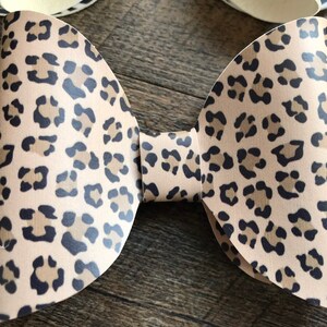 May include: A tan and black leopard print bow. The bow is made of a flexible material and has a classic bow shape. The leopard print features dark brown and black spots on a tan background. Other animal print bows are visible.