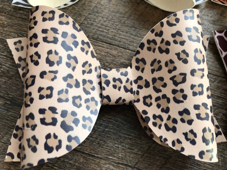 May include: A tan and black leopard print bow. The bow is made of a flexible material and has a classic bow shape. The leopard print features dark brown and black spots on a tan background. Other animal print bows are visible.