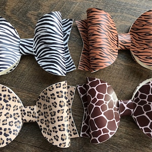 May include: Four large hair bows with animal print patterns. The bows feature zebra stripes, tiger stripes, leopard spots, and giraffe spots. The bows are made of a shiny material and are arranged on a wooden surface.