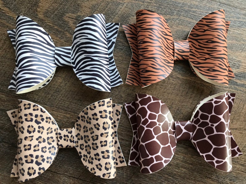 May include: Four large hair bows with animal print patterns. The bows feature zebra stripes, tiger stripes, leopard spots, and giraffe spots. The bows are made of a shiny material and are arranged on a wooden surface.