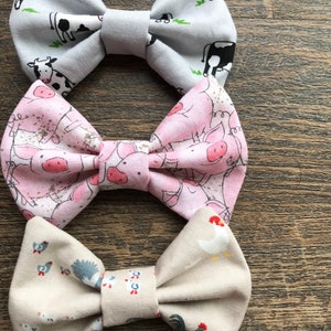 May include: Three fabric bow ties with farm animal prints. The top bow tie is gray with black and white cows, the middle bow tie is pink with pink pigs, and the bottom bow tie is beige with white chickens.