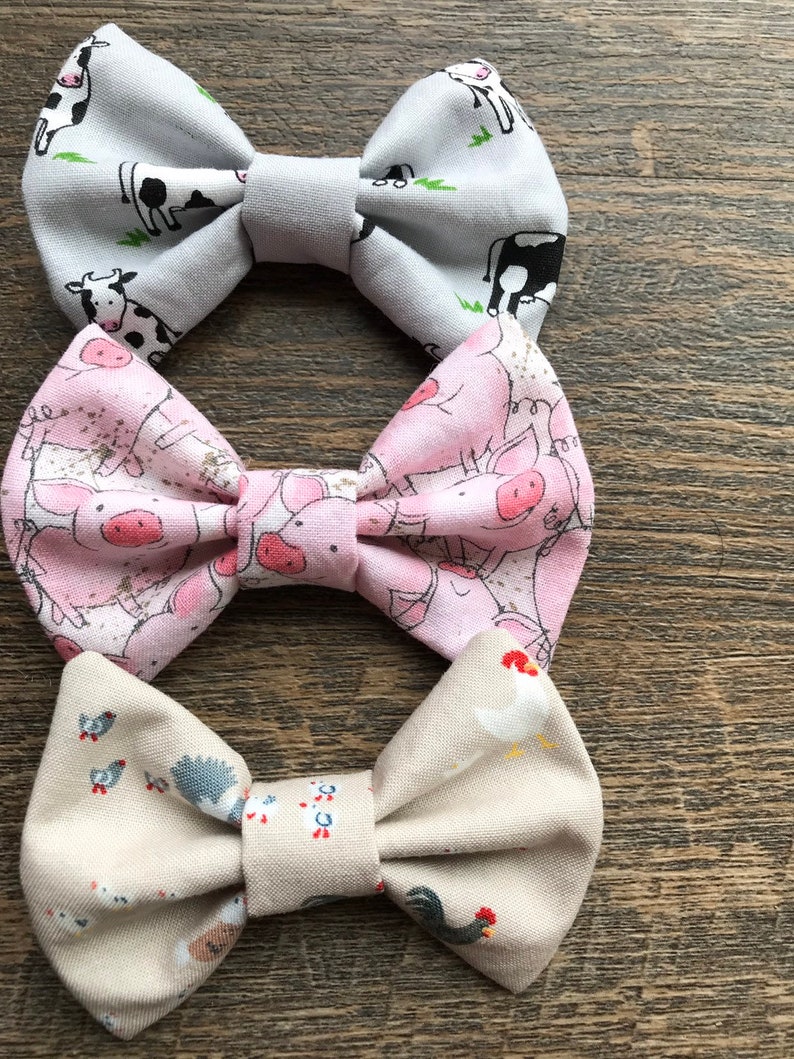 May include: Three fabric bow ties with farm animal prints. The top bow tie is gray with black and white cows, the middle bow tie is pink with pink pigs, and the bottom bow tie is beige with white chickens.
