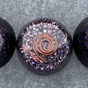May include: Three purple and copper orgonite pyramids with a copper coil inside each. The pyramids are on a grey surface.