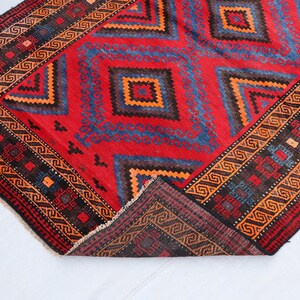 Pode incluir: Um tapete vermelho com um padrão geométrico de losangos. O tapete apresenta um campo central vermelho com formas de losangos azuis, laranja e pretos. A borda é marrom escuro com padrões geométricos. O tapete está parcialmente dobrado, revelando o verso.