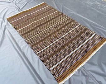 Puede incluir: Una alfombra tejida rectangular con un patrón repetitivo de rayas horizontales en tonos amarillo mostaza, marrón oscuro, blanco y rojo intenso. El diseño incluye motivos geométricos complejos en las bandas y flecos blancos en los extremos.
