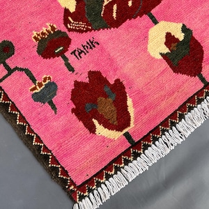 K&ouml;nnte beinhalten: Rosa Teppich mit einem floralen Design in Burgunderrot, Creme und Dunkelgr&uuml;n. Der Teppich hat einen dunkelbraunen Rand mit Zickzackmuster und wei&szlig;en Fransen. Das Wort "TANK" ist auf dem Teppich zu sehen.