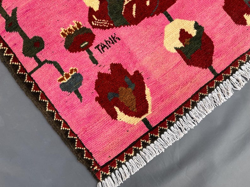 K&ouml;nnte beinhalten: Rosa Teppich mit einem floralen Design in Burgunderrot, Creme und Dunkelgr&uuml;n. Der Teppich hat einen dunkelbraunen Rand mit Zickzackmuster und wei&szlig;en Fransen. Das Wort "TANK" ist auf dem Teppich zu sehen.