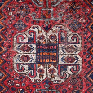 Pu&ograve; includere: Un tappeto vintage con sfondo rosso e intricati motivi geometrici in tonalit&agrave; di blu, bianco e arancio. Il tappeto ha un aspetto usurato e sbiadito, che gli conferisce un aspetto rustico e antico.