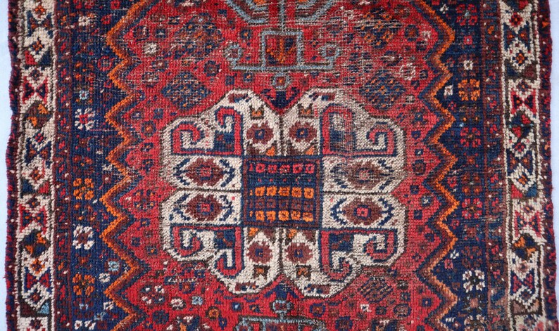Pu&ograve; includere: Un tappeto vintage con sfondo rosso e intricati motivi geometrici in tonalit&agrave; di blu, bianco e arancio. Il tappeto ha un aspetto usurato e sbiadito, che gli conferisce un aspetto rustico e antico.