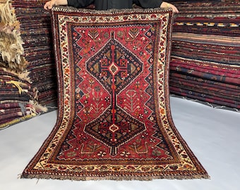 May include: A rectangular hand-knotted rug featuring a deep red field with a large central dark blue diamond medallion. The rug is decorated with intricate geometric and floral motifs in cream, orange, and black, finished with a fringed edge.