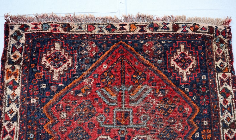 Pu&ograve; includere: Un tappeto persiano vintage con uno schema di colori rosso, blu e crema. Il tappeto ha una forma di diamante centrale con intricati motivi floreali e un bordo con disegni geometrici.