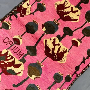 K&ouml;nnte beinhalten: Rosa Teppich mit einem sich wiederholenden Blumenmuster in Creme, Burgunderrot und Dunkelgrau. Das Wort "OPIUM" ist in wei&szlig;er Schrift zu sehen. Der Teppich hat einen dunkelbraunen Rand und misst ca. 150 cm x 90 cm.