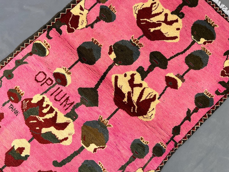 K&ouml;nnte beinhalten: Rosa Teppich mit einem sich wiederholenden Blumenmuster in Creme, Burgunderrot und Dunkelgrau. Das Wort "OPIUM" ist in wei&szlig;er Schrift zu sehen. Der Teppich hat einen dunkelbraunen Rand und misst ca. 150 cm x 90 cm.