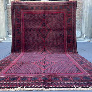 Può includere: Un grande tappeto rettangolare con un motivo geometrico bordeaux scuro e nero. Il tappeto presenta un disegno a diamante centrale e un bordo dettagliato. Il tappeto è esposto all'aperto, mostrando le sue dimensioni e i suoi dettagli intricati.