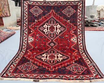 Puede incluir: Una alfombra roja rectangular grande con un diseño geométrico central en blanco y negro. La alfombra presenta un patrón de diamantes repetido y un borde blanco con detalles intrincados. La alfombra se exhibe al aire libre.