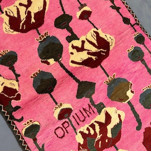 K&ouml;nnte beinhalten: Ein pinkfarbener Teppich mit einem sich wiederholenden Muster aus stilisierten Blumen und Samenkapseln in Creme, Burgunderrot und Dunkelgr&uuml;n. Das Wort "OPIUM" ist im Design sichtbar. Der Teppich hat einen dunklen Rand.