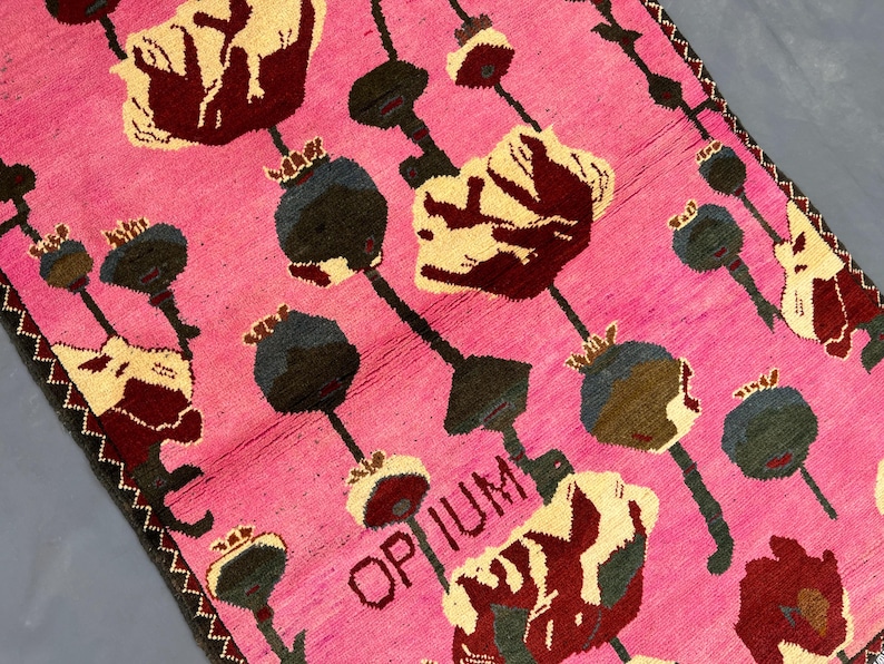 K&ouml;nnte beinhalten: Ein pinkfarbener Teppich mit einem sich wiederholenden Muster aus stilisierten Blumen und Samenkapseln in Creme, Burgunderrot und Dunkelgr&uuml;n. Das Wort "OPIUM" ist im Design sichtbar. Der Teppich hat einen dunklen Rand.