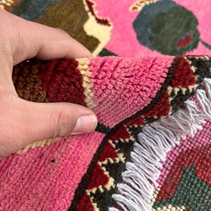 K&ouml;nnte beinhalten: Nahaufnahme eines Teils eines rosa Teppichs mit floralem Design. Der Teppich weist eine Kombination aus rosa, roten, gr&uuml;nen und cremefarbenen Farben auf. Der Rand des Teppichs hat eine dekorative Bord&uuml;re und einen wei&szlig;en Fransen.