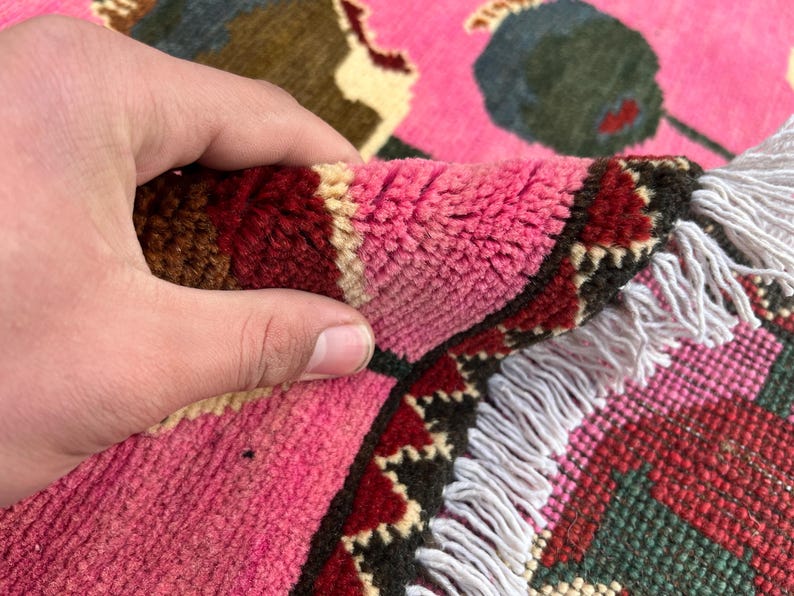 K&ouml;nnte beinhalten: Nahaufnahme eines Teils eines rosa Teppichs mit floralem Design. Der Teppich weist eine Kombination aus rosa, roten, gr&uuml;nen und cremefarbenen Farben auf. Der Rand des Teppichs hat eine dekorative Bord&uuml;re und einen wei&szlig;en Fransen.