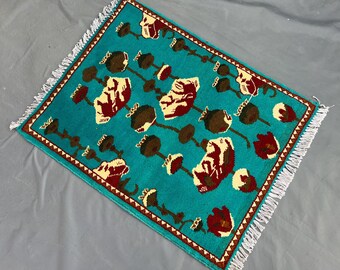 Puede incluir: Alfombra rectangular turquesa con un patrón floral repetido en marrón y crema. Tiene un borde marrón y flecos blancos. El diseño sugiere una fabricación artesanal, con patrones detallados.