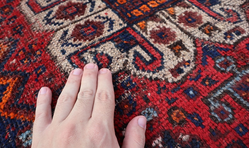 Pu&ograve; includere: Primo piano di un tappeto rosso, blu e bianco con un motivo con una mano appoggiata sulla superficie.