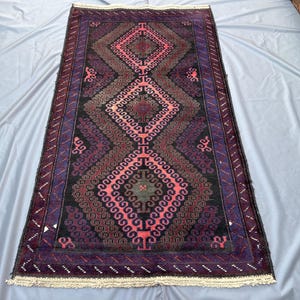 Pode incluir: Um tapete retangular com um padrão de diamante central em tons de vermelho, rosa e preto. O tapete tem uma borda roxa escura com um design geométrico repetitivo. O design do tapete é simétrico e intrincado, com franjas nas extremidades curtas.