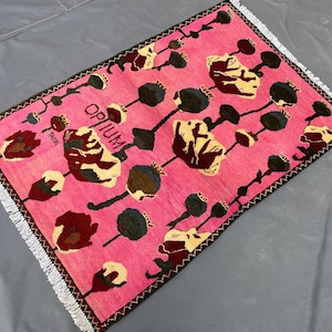 K&ouml;nnte beinhalten: Ein rechteckiger, pinkfarbener Teppich mit floralem Muster und dem Wort "OPIUM" in Schwarz. Der Teppich hat einen dunkelbraunen Rand und wei&szlig;e Fransen. Das florale Design umfasst beige, dunkelrote und schwarze Elemente.