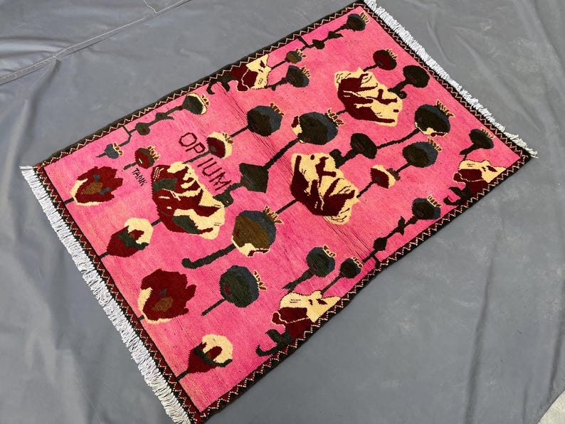 K&ouml;nnte beinhalten: Ein rechteckiger, pinkfarbener Teppich mit floralem Muster und dem Wort "OPIUM" in Schwarz. Der Teppich hat einen dunkelbraunen Rand und wei&szlig;e Fransen. Das florale Design umfasst beige, dunkelrote und schwarze Elemente.