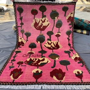 K&ouml;nnte beinhalten: Ein rechteckiger, pinkfarbener Teppich mit einem floralen Muster aus roten, cremefarbenen und gr&uuml;nen mohnartigen Bl&uuml;ten und Samenkapseln. Der Teppich hat einen schwarzen Rand und wei&szlig;en Fransen. Das Wort "OPIUM" ist auf dem Teppich aufgedruckt.