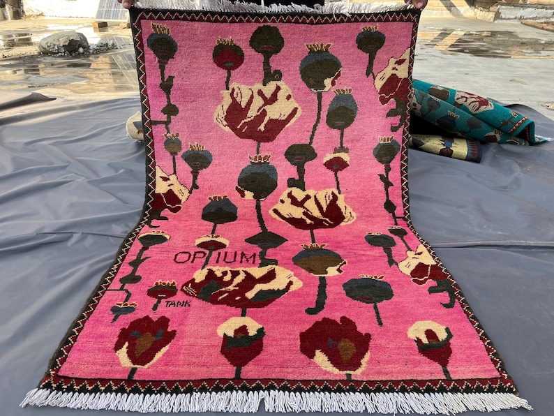 K&ouml;nnte beinhalten: Ein rechteckiger, pinkfarbener Teppich mit einem floralen Muster aus roten, cremefarbenen und gr&uuml;nen mohnartigen Bl&uuml;ten und Samenkapseln. Der Teppich hat einen schwarzen Rand und wei&szlig;en Fransen. Das Wort "OPIUM" ist auf dem Teppich aufgedruckt.