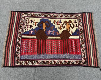 Puede incluir: Una alfombra tradicional con un patrón geométrico rojo, azul y blanco. Dos carneros marrones con cuernos curvos se representan en el centro de la alfombra.